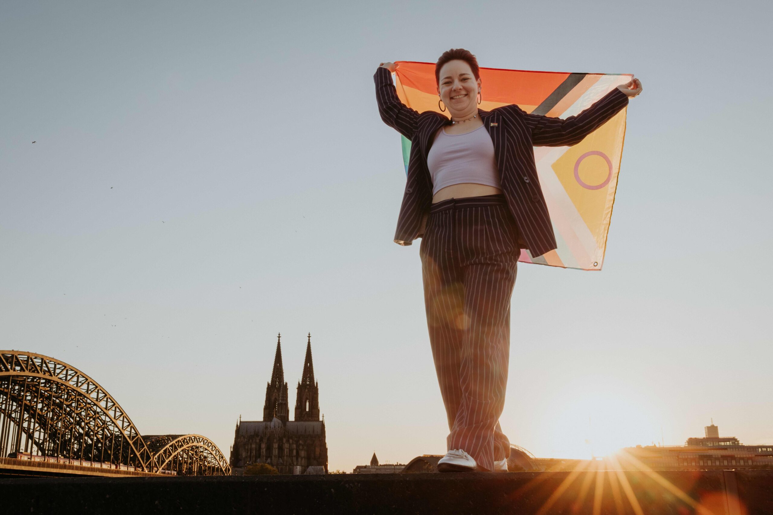 Freie Rednerin Caro steht am Rhein mit Regenbogenflagge in der Hand und Blick auf den Kölner Dom